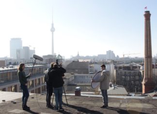 Im Vordergrund dreht eine 5-köpfige Gruppe auf einem Dach einen Film. Links hält eine Person ein großes Mikro, eine andere Person rechts einen Reflektor, in der Mitte stehen zwei Personen hinter und eine vor der Kamera. Im Hintergrund befindet sich die Stadtsilhouette Berlins einschließlich des Fernsehturms an einem sonnigen, wolkenlosen Tag.