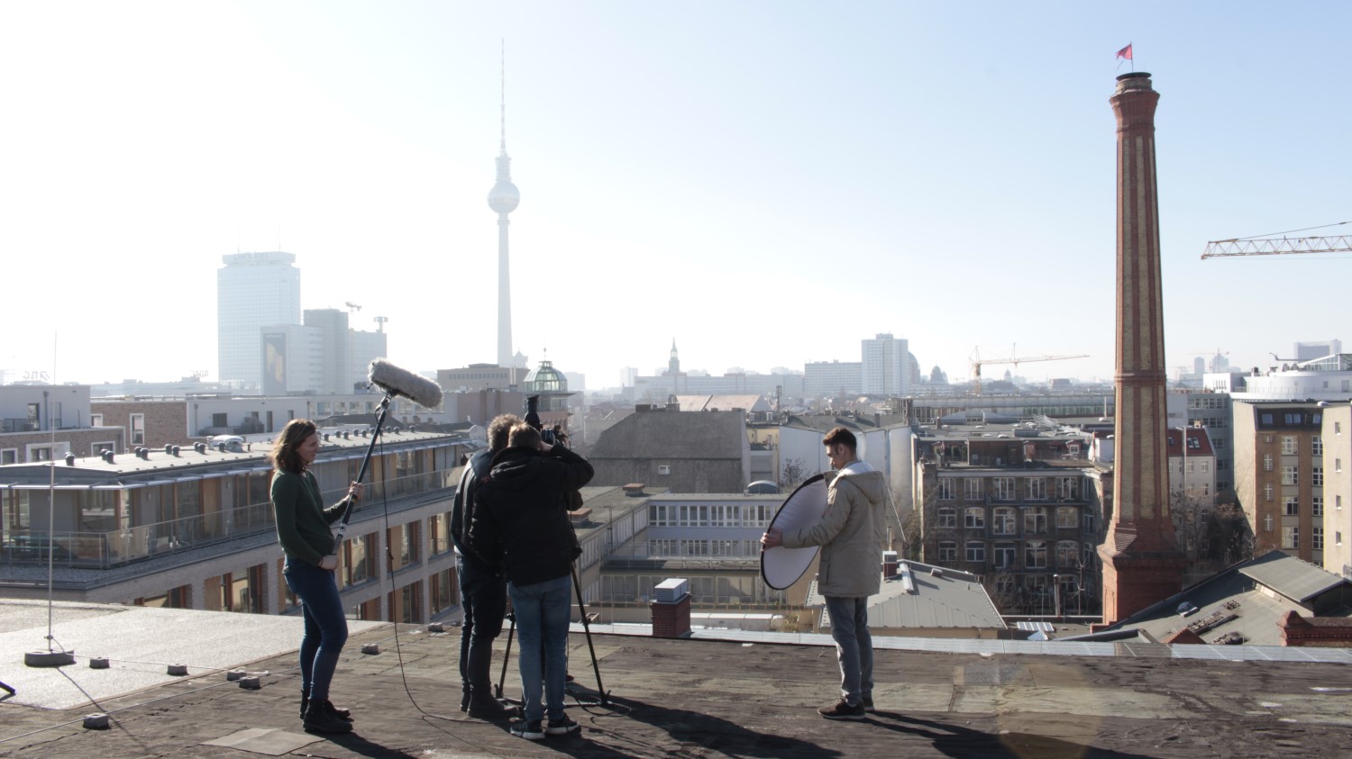 Im Vordergrund dreht eine 5-köpfige Gruppe auf einem Dach einen Film. Links hält eine Person ein großes Mikro, eine andere Person rechts einen Reflektor, in der Mitte stehen zwei Personen hinter und eine vor der Kamera. Im Hintergrund befindet sich die Stadtsilhouette Berlins einschließlich des Fernsehturms an einem sonnigen, wolkenlosen Tag.