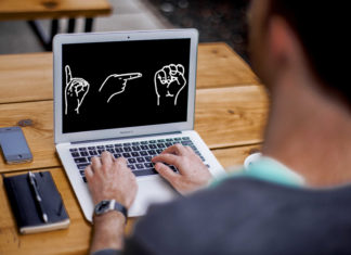 Eine Person sitzt vor einem Laptop, auf dem "DGS" im Fingeralphabet abgebildet ist.