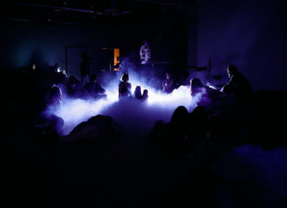 Das Bild zeigt eine Szene aus der Inszenierung Herbarium aus dem Jahr 2022. Das Bild ist dunkel. In der Mitte der Szene steigt Nebel auf. Der Nebel ist blau ausgeleuchtet. Im Nebel und um den Nebel herum befinden sich Menschen, deren Silhouetten zu sehen sind.