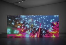 Eine große Leinwand steht in einem abgedunkelten Raum. Auf der Leinwand sind ganz viele weiße Punkte und grelle Farben zu sehen.