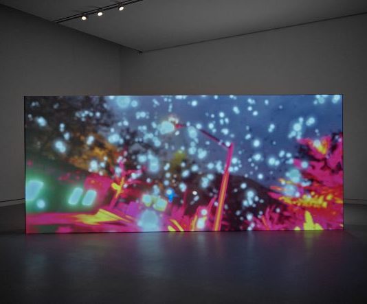 Eine große Leinwand steht in einem abgedunkelten Raum. Auf der Leinwand sind ganz viele weiße Punkte und grelle Farben zu sehen.