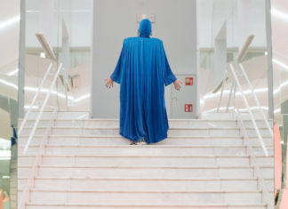 Eine weiße Treppe, die links und rechts von verspiegelten Wänden eingegrenzt wird. Auf dessen Mitte steht eine Person. Sie ist von hinten zu sehen und steckt von Kopf bis Fuß in einem langärmeligen, fließenden Gewand in der Farbe Blau. Beide Arme sind leicht ausgestreckt, die offenen Handinnenflächen zeigen in die Richtung des weißen Raums, zu dem die Treppe führt.