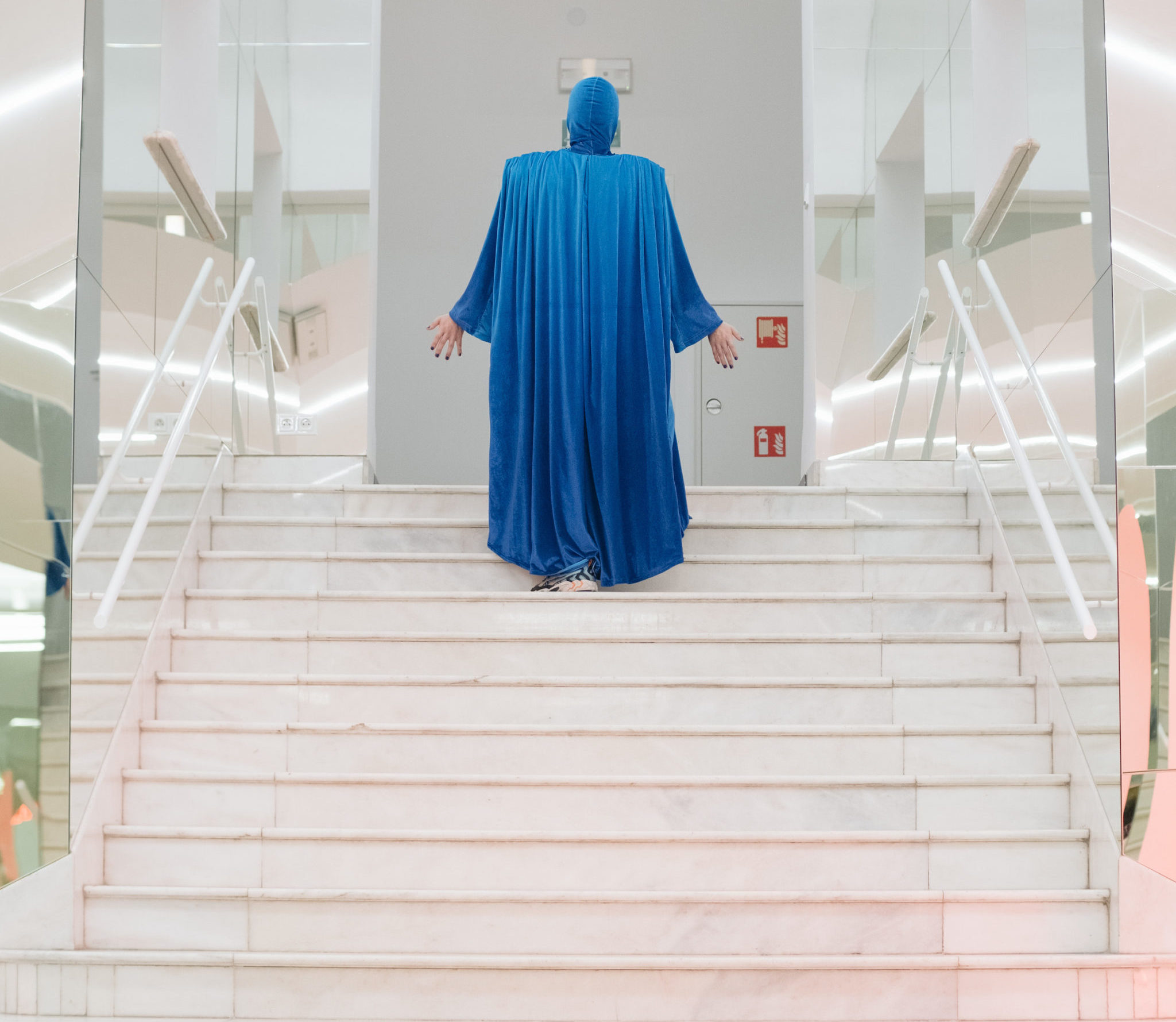 Eine weiße Treppe, die links und rechts von verspiegelten Wänden eingegrenzt wird. Auf dessen Mitte steht eine Person. Sie ist von hinten zu sehen und steckt von Kopf bis Fuß in einem langärmeligen, fließenden Gewand in der Farbe Blau. Beide Arme sind leicht ausgestreckt, die offenen Handinnenflächen zeigen in die Richtung des weißen Raums, zu dem die Treppe führt.