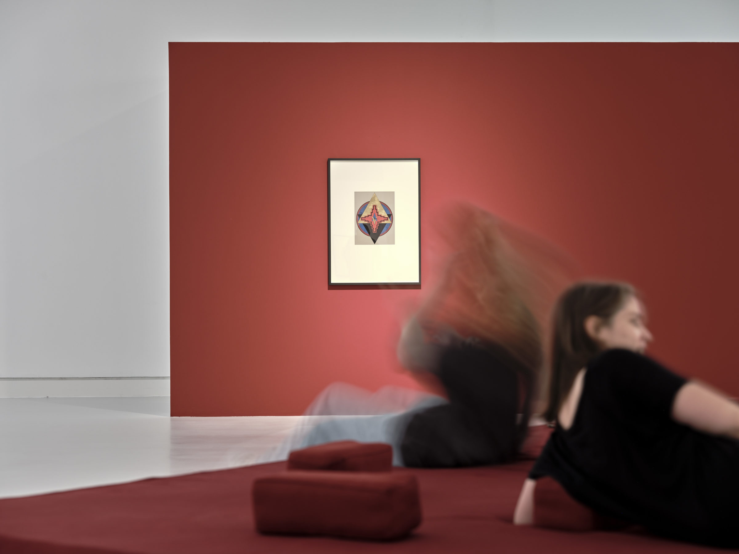 An einer roten Wand hängt ein Bild. Auf dem Bild sind geometrische Formen. Vor der Wand steht eine rote Sitzgelegenheit. Darauf sitzen zwei Personen.