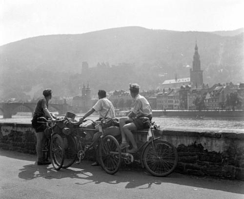 Auf einer schwarz-weiß Fotografie sitzen drei junge Männer auf der Ufermauer des Neckars. Ihre Fahrräder stehen vor ihnen und ihre Blicke gehen in Richtung Heidelberger Schloss, das im Hintergrund vor der Bergsilhouette zu sehen ist.