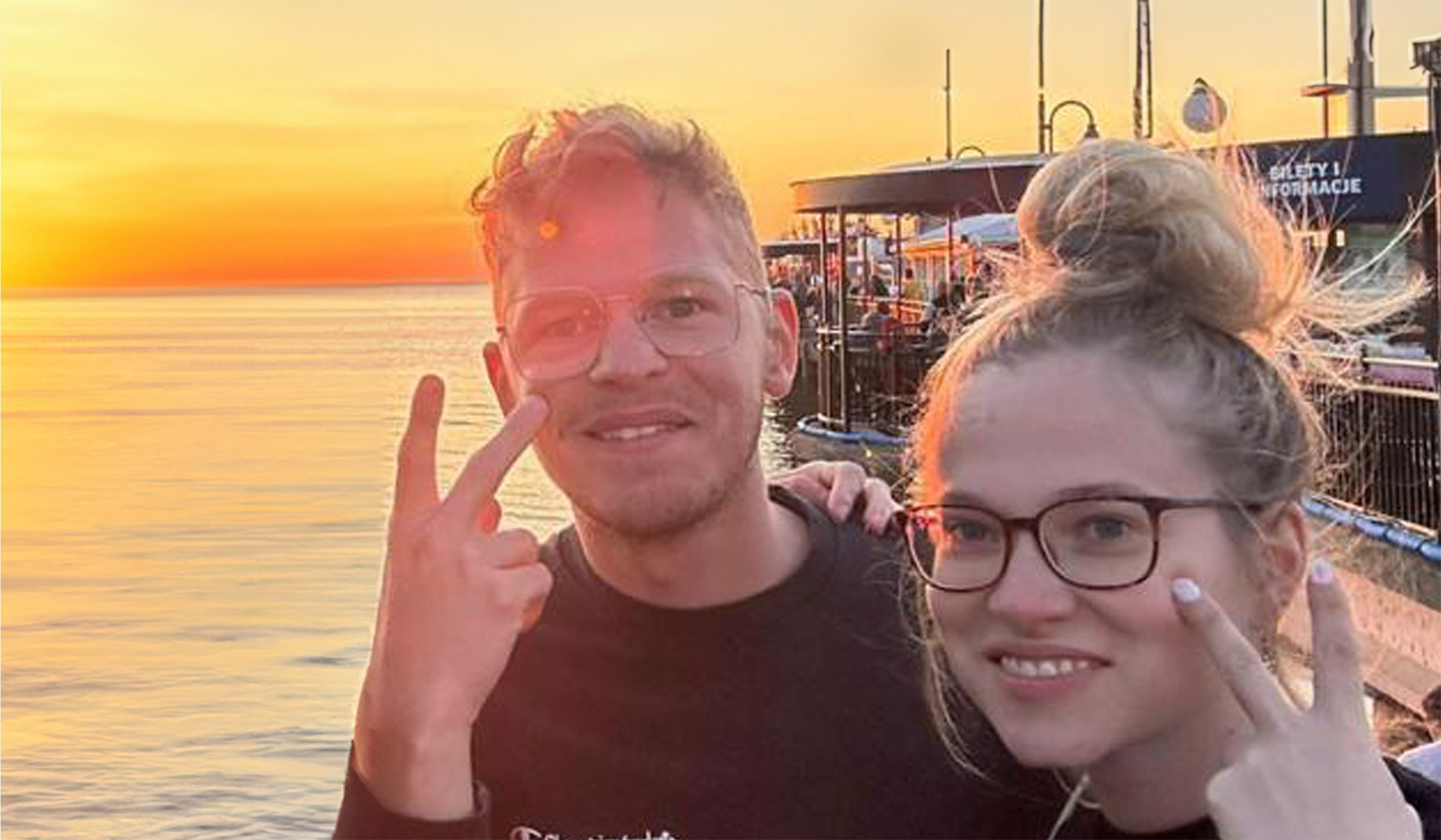 Ein Foto von Marcel Fischer und Lore Bajerski. Links im Bild ist Marcel Fischer. Er trägt eine Brille mit dünner goldener Fassung, hat kurze blonde lockige Haare, einen Dreitagebart und trägt einen schwarzen Pullover. Neben ihm, rechts im Bild, steht Lore Bajerski. Sie hat ihre blonden Haare in einem hohen Dutt zusammengebunden, trägt eine Brille mit dunkler Fassung, lackierte Fingernägel, eine Kette und ebenfalls einen schwarzen Pullover. Beide lächeln in die Kamera und halten jeweils den Mittelfinger einer Hand in V-Handform an ihre Wange. Der Hintergrund zeigt die Abendsonne über dem Meer und rechts im Bild sind Stände am Pier zu erkennen