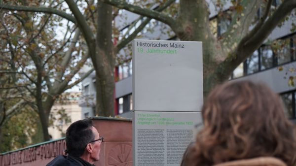 Auf dem Bild ist eine Säule zu sehen. Auf der Säule steht "Historisches Mainz. 19. Jahrhundert. Vor der Säule stehen zwei Menschen. Man sieht die Hinterköpfe der zwei Menschen.