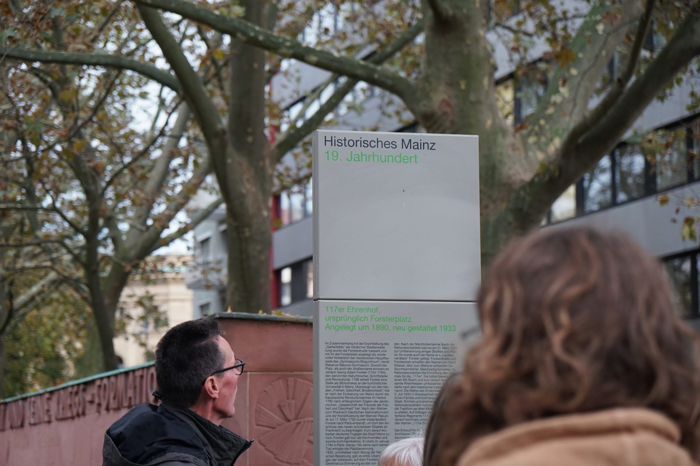 Auf dem Bild ist eine Säule zu sehen. Auf der Säule steht "Historisches Mainz. 19. Jahrhundert. Vor der Säule stehen zwei Menschen. Man sieht die Hinterköpfe der zwei Menschen.