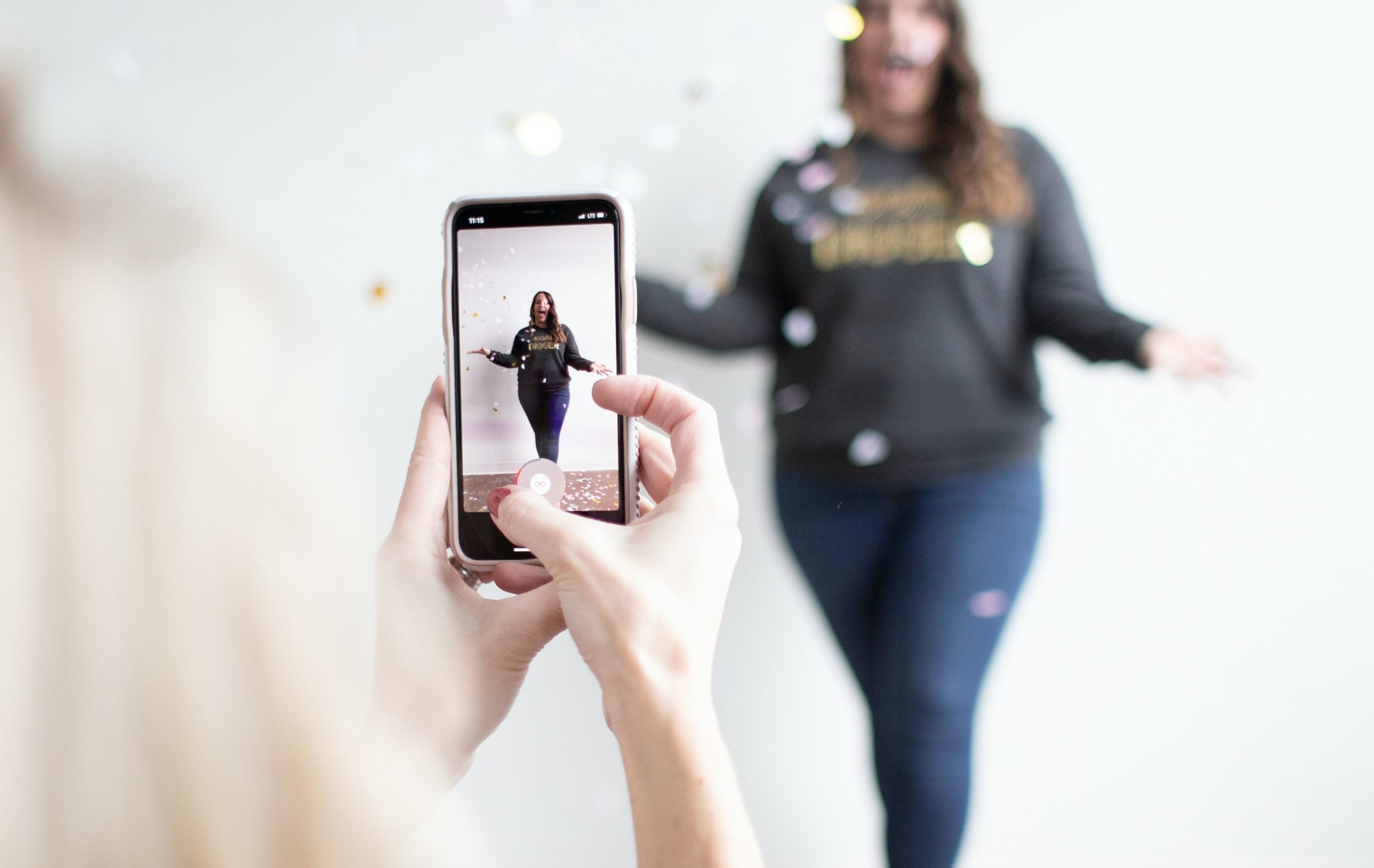 Sicht auf eine blonde Person, die mit dem Smartphone eine weiblich-gelesene brünette Person im Hintergrund dabei filmt, wie diese Konfetti wirft.