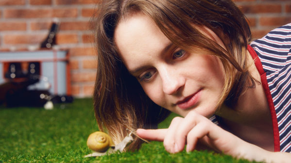 Nahaufnahme einer jungen Frau, die auf dem Bauch auf dem Rasen liegt und mit einer Schnecke spielt. Im Hintergrund eine Backsteinmauer und ein Bierkasten mit leeren Flaschen.,