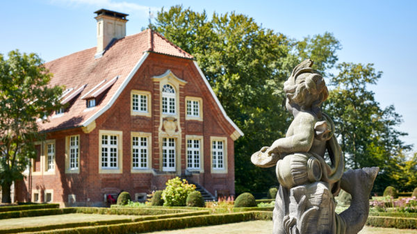 Das Bild zeigt das Haus Rüschhaus - ein denkmalgeschütztes, barockes Gebäude, das links angeschnitten ist. Im rechten Vordergrund befindet sich eine Sandsteinskultpur in Form eines Puttos, dessen Blick auf das Gebäude gerichtet ist. Der Himmel ist blau, hinter dem Gebäude erstreckt sich das Grün von Bäumen. Zwischen Gebäude und Putto sieht man ein Stück des Gartens mit niedriger Buchsbaumhecke sonnenverbranntem Rasen.