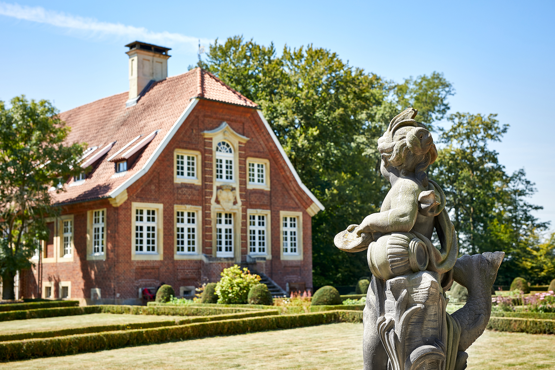 Das Bild zeigt das Haus Rüschhaus - ein denkmalgeschütztes, barockes Gebäude, das links angeschnitten ist. Im rechten Vordergrund befindet sich eine Sandsteinskultpur in Form eines Puttos, dessen Blick auf das Gebäude gerichtet ist. Der Himmel ist blau, hinter dem Gebäude erstreckt sich das Grün von Bäumen. Zwischen Gebäude und Putto sieht man ein Stück des Gartens mit niedriger Buchsbaumhecke sonnenverbranntem Rasen.