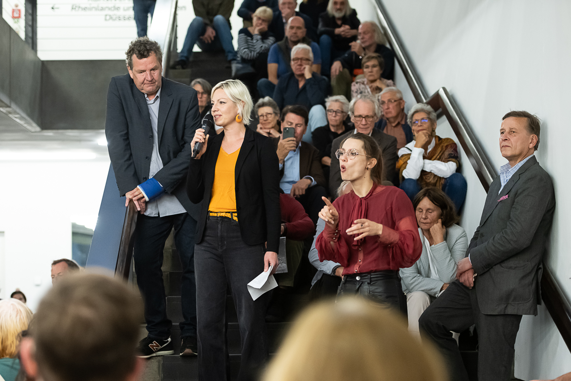 Fotografie von einer Gruppe Menschen, die auf einer Treppe sitzt. Vor ihnen stehen drei Personen, ein Mann und zwei Frauen. Die Frau in der Mitte spricht durch ein Mikrophon, die Frau rechts verdolmetscht das Gesagte in DGS.