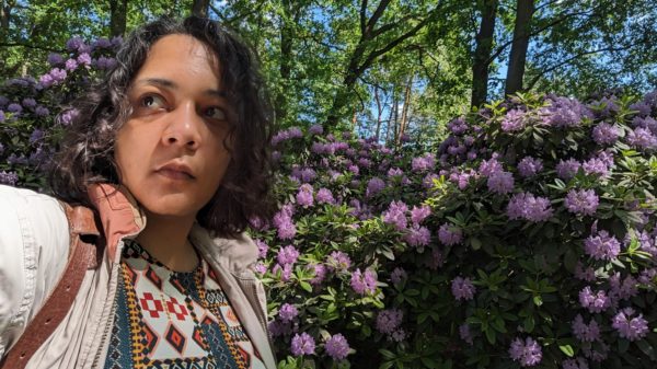 Eine Frau mit dunklem lockigen Haar schaut aus dem Bild nach links. Rechts neben ihr ist ein Busch mit lilanen Blüten