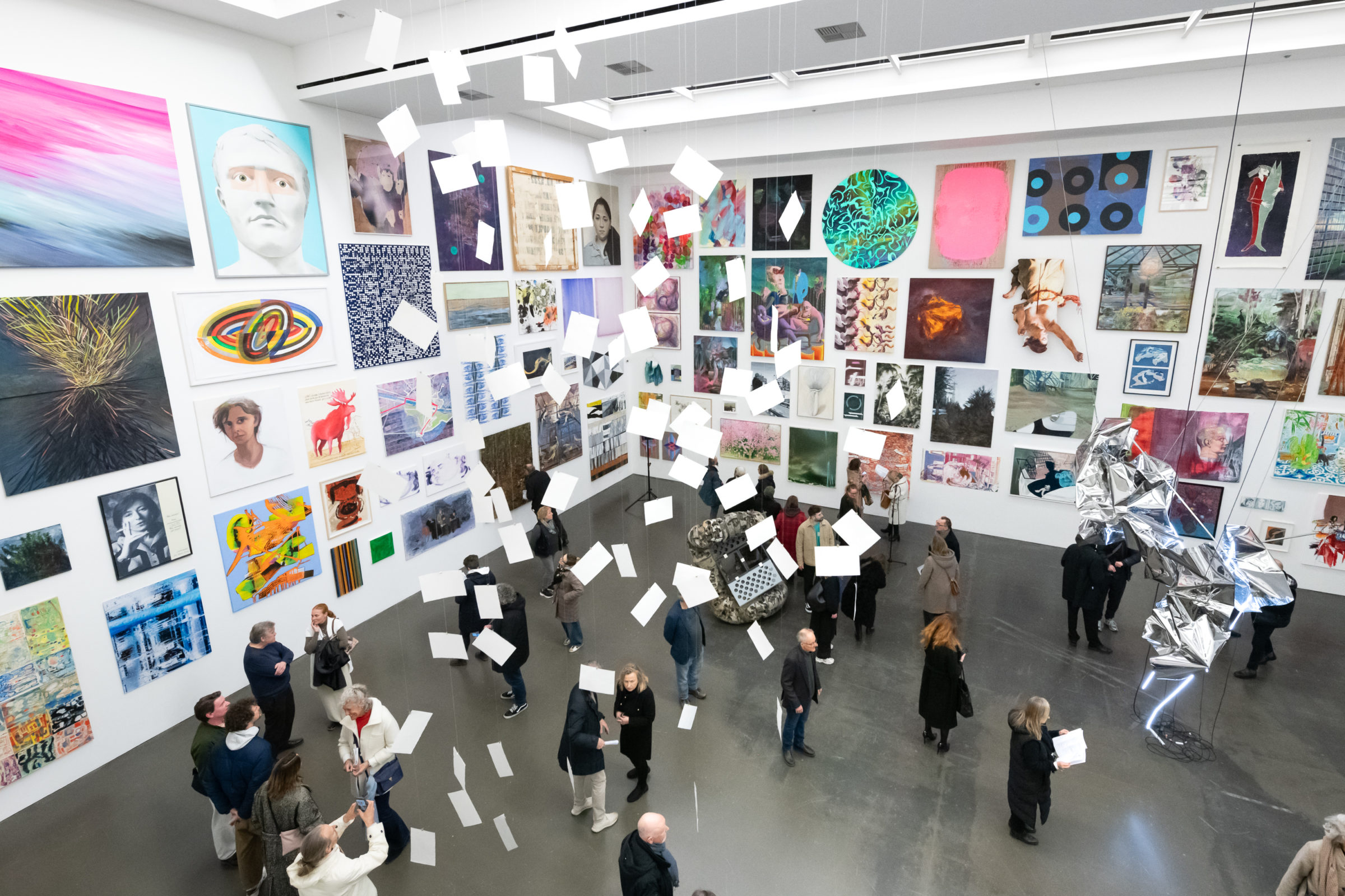 Ein heller Ausstellungsraum mit hohen weißen Wänden, dicht behängt mit zahlreichen Kunstwerken in verschiedenen Stilen und Formaten. In der Mitte des Raums schweben weiße Papierblätter, die von der Decke herabhängen. Besucherinnen und Besucher bewegen sich durch den Raum, betrachten die Werke, unterhalten sich oder machen Fotos. Rechts im Bild steht eine glänzende, skulpturale Installation mit geometrischen Formen und Lichtelementen. Die Atmosphäre wirkt lebendig und interessiert.