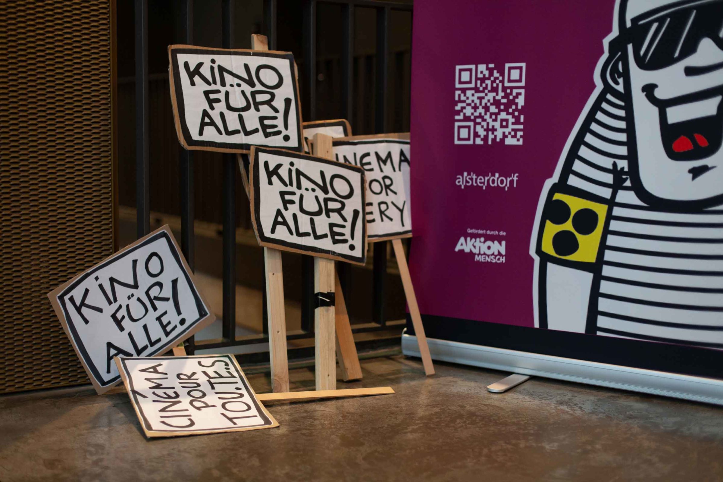 Mehrere Schilder mit der Aufschrift "Kino für Alle" stehen und liegen an einem Geländer.