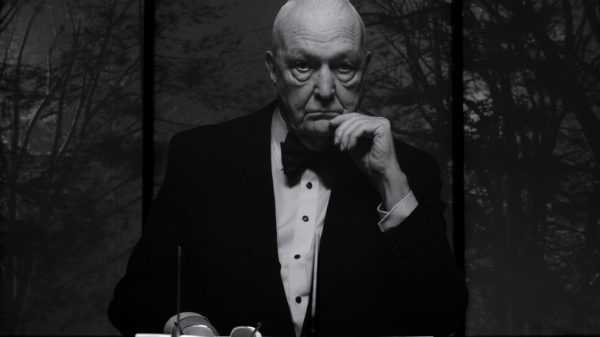 Das Bild zeigt einen älteren Mann in einem eleganten schwarzen Anzug mit Fliege. Sein Gesichtsausdruck ist ernst und nachdenklich. Er stützt sein Kinn auf die Hand und blickt direkt in die Kamera. Das Bild zeigt einen älteren Mann in einem eleganten schwarzen Anzug mit Fliege. Sein Gesichtsausdruck ist ernst und nachdenklich. Er stützt sein Kinn auf die Hand und blickt direkt in die Kamera.