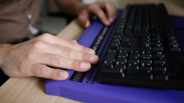 Eine Person nutzt eine Braillezeile vor einer herkömmlichen schwarzen Tastatur. Die linke Hand tastet die erhabenen Braille-Zeichen, während die rechte Hand auf der Tastatur ruht. Die Braillezeile ist lila und liegt auf einem Holztisch.