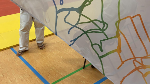 Foto: Bewegte weiße Fahne mit blauen, grünen, orangen Figurenzeichnungen im Vordergrund; dahinter: Beine eines Judoka im weißen Judoanzug, der auf einem hölzernen Turnhallenboden steht.