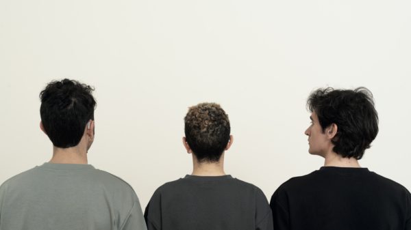 Drei Personen stehen mit dem Rücken zur Kamera vor einer weißen Wand. Die Person in der Mitte hat lockiges Haar mit helleren Spitzen, die beiden anderen haben dunkles, glattes Haar. Zwei der Personen tragen sichtbare Hörgeräte.