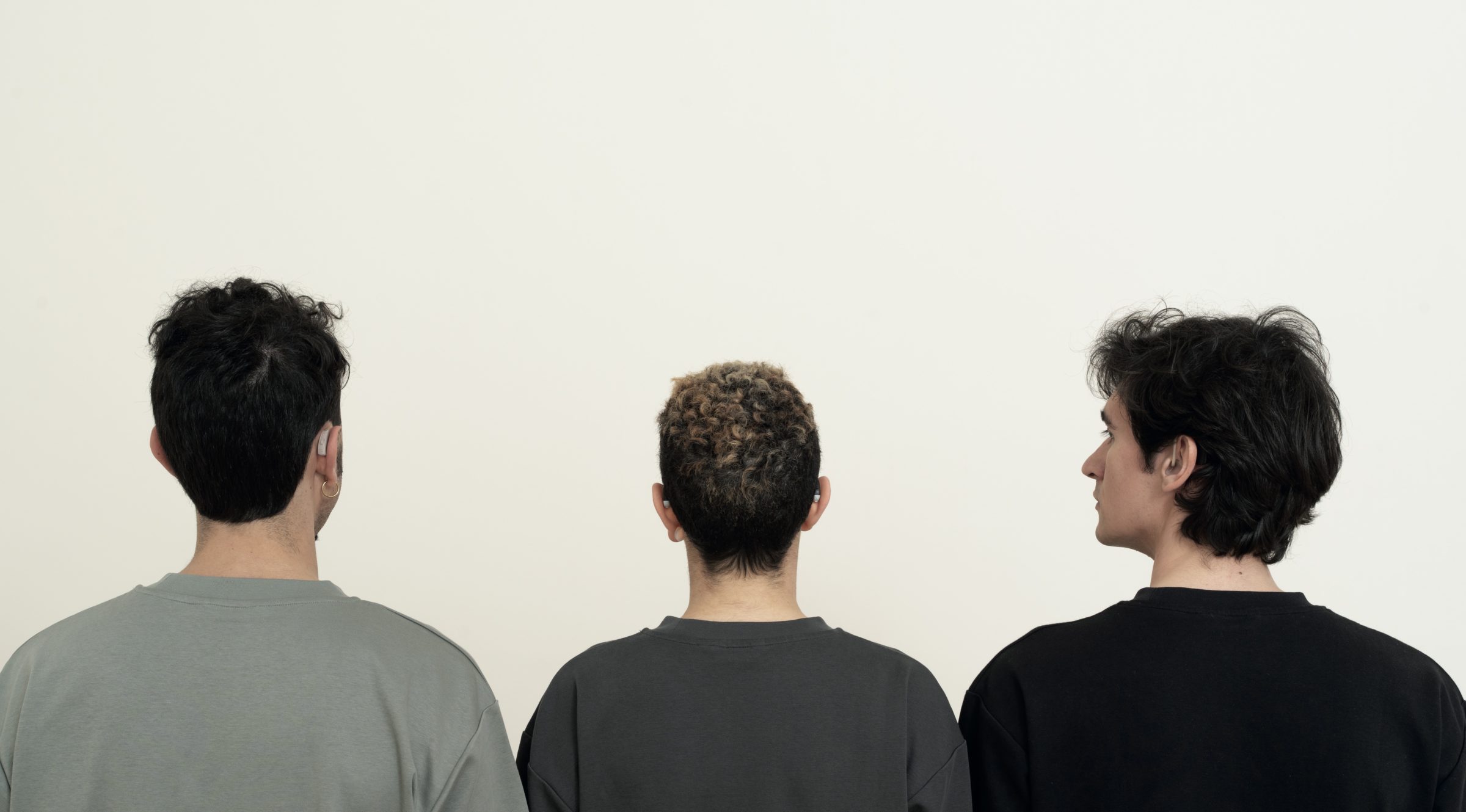 Drei Personen stehen mit dem Rücken zur Kamera vor einer weißen Wand. Die Person in der Mitte hat lockiges Haar mit helleren Spitzen, die beiden anderen haben dunkles, glattes Haar. Zwei der Personen tragen sichtbare Hörgeräte.