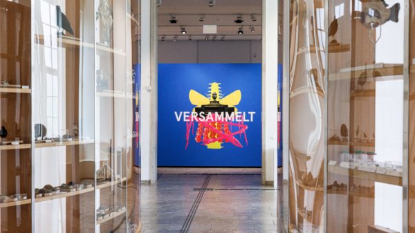 Foto: Blick zur Ausstellung Versammelt. Ein taktiles Bodenleitsystem führt zwischen zwei Regalwänden durch auf eine blaue Wand zu. Auf der Wand steht in weiß "Versammelt".
