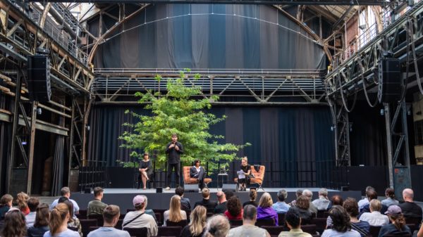 Veranstaltung der Diskursreihe Brave New Voices in der Jahrhunderthalle Bochum. Im Vordergrund ist Publikum von hinten zu erkennen, im Hintergrund zwei Personen auf einer Bühne sitzend und sich austauschend.