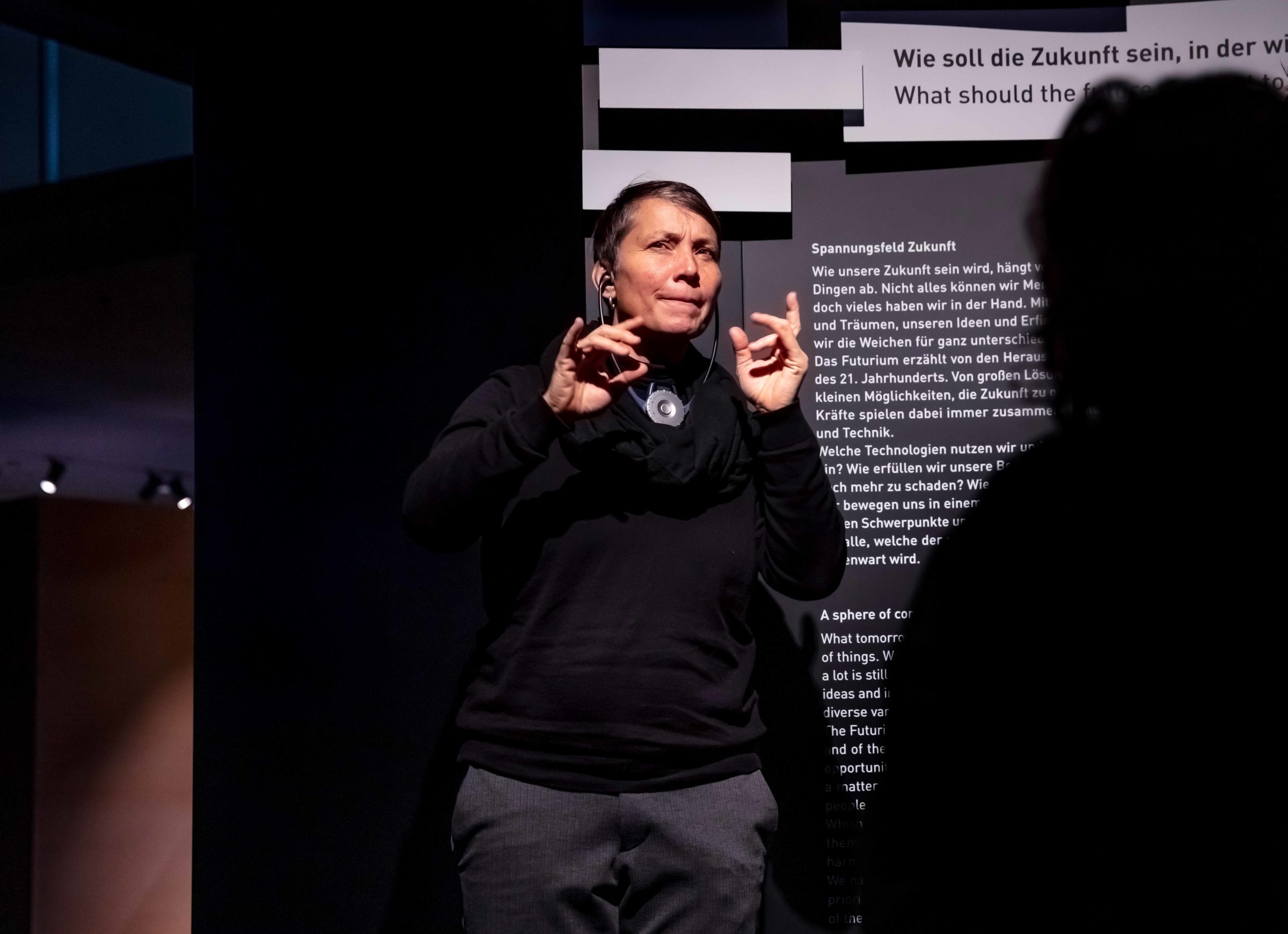 Dunkel bekleidete Frau spricht in Gebärdensprache. Im Hintergrund ist ein Museumsaufsteller zu sehen.