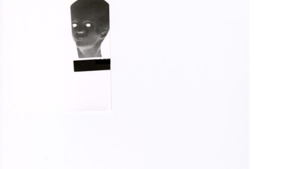 Das Bild zeigt einen weißen Hintergrund mit einer kleinen schwarz-weißen Figur im Negativstil am linken Rand. Die Figur stellt einen Kopf mit leuchtend weißen Augen dar. Direkt ober- und unterhalb des Kopfes befinden sich zwei rechteckige schwarze Balken. Außerdem ist erkennbar, dass das Motiv auf einem separaten Stück Papier gedruckt ist, das wiederum auf einem weißen Blatt Papier liegt. Das Bild zeigt einen weißen Hintergrund mit einer kleinen schwarz-weißen Figur im Negativstil am linken Rand. Die Figur stellt einen Kopf mit leuchtend weißen Augen dar. Direkt ober- und unterhalb des Kopfes befinden sich zwei rechteckige schwarze Balken. Außerdem ist erkennbar, dass das Motiv auf einem separaten Stück Papier gedruckt ist, das wiederum auf einem weißen Blatt Papier liegt.