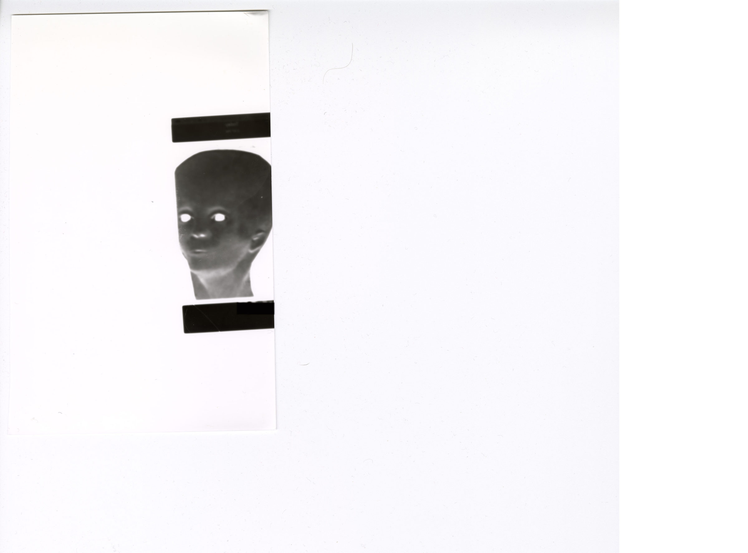 Das Bild zeigt einen weißen Hintergrund mit einer kleinen schwarz-weißen Figur im Negativstil am linken Rand. Die Figur stellt einen Kopf mit leuchtend weißen Augen dar. Direkt ober- und unterhalb des Kopfes befinden sich zwei rechteckige schwarze Balken. Außerdem ist erkennbar, dass das Motiv auf einem separaten Stück Papier gedruckt ist, das wiederum auf einem weißen Blatt Papier liegt.