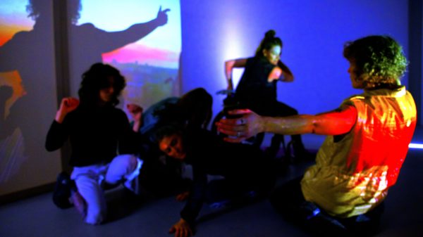 In einem zeitlosen, blau beleuchteten Raum bewegt sich eine Gruppe von fünf weißen Körpern vor einem projizierten Sonnenuntergang. Das Licht spiegelt sich auf ihrer Haut und wirft leuchtend rote, blaue und orangefarbene Streifen. In der Mitte schmelzen zwei Körper in Richtung Boden, verlagern ihr Gewicht und geben sich der Schwerkraft hin. Unten trifft eine weiche, offene Handfläche auf den Boden, trotzt dem Zusammenbruch, der Kopf ist leicht geneigt und ruht auf einem Luftkissen. Zwei umkreisende Körper beziehen ihre Energie aus dem Kern, ihre Präsenz ist aufgeladen. Die Anziehungskraft bestimmt ihre Bewegungen: ein nach oben gebeugter Arm, eine Hand, die sich am Rollstuhl festhält und freudig eine Drehung erwartet; ein weiteres Paar Arme, die sich erhoben haben, weiche, geschlossene Fäuste, die entschlossen in der Luft schweben. Mit dem Gesicht zur Leinwand umarmt eine Person mit lockigem Haar den Raum um die Gruppe herum. Mit ausgestreckten Armen kniet sie zurück, stark und fürsorglich, und dehnt dessen Grenzen aus. Ihr Rücken wird nun zu einer großen roten Sonne, die auf eine goldene, ärmellose Weste aus den 80er Jahren scheint. Ein Schatten fällt auf die gesättigte Projektion und verdoppelt die Sonnenfigur über der Landschaft.