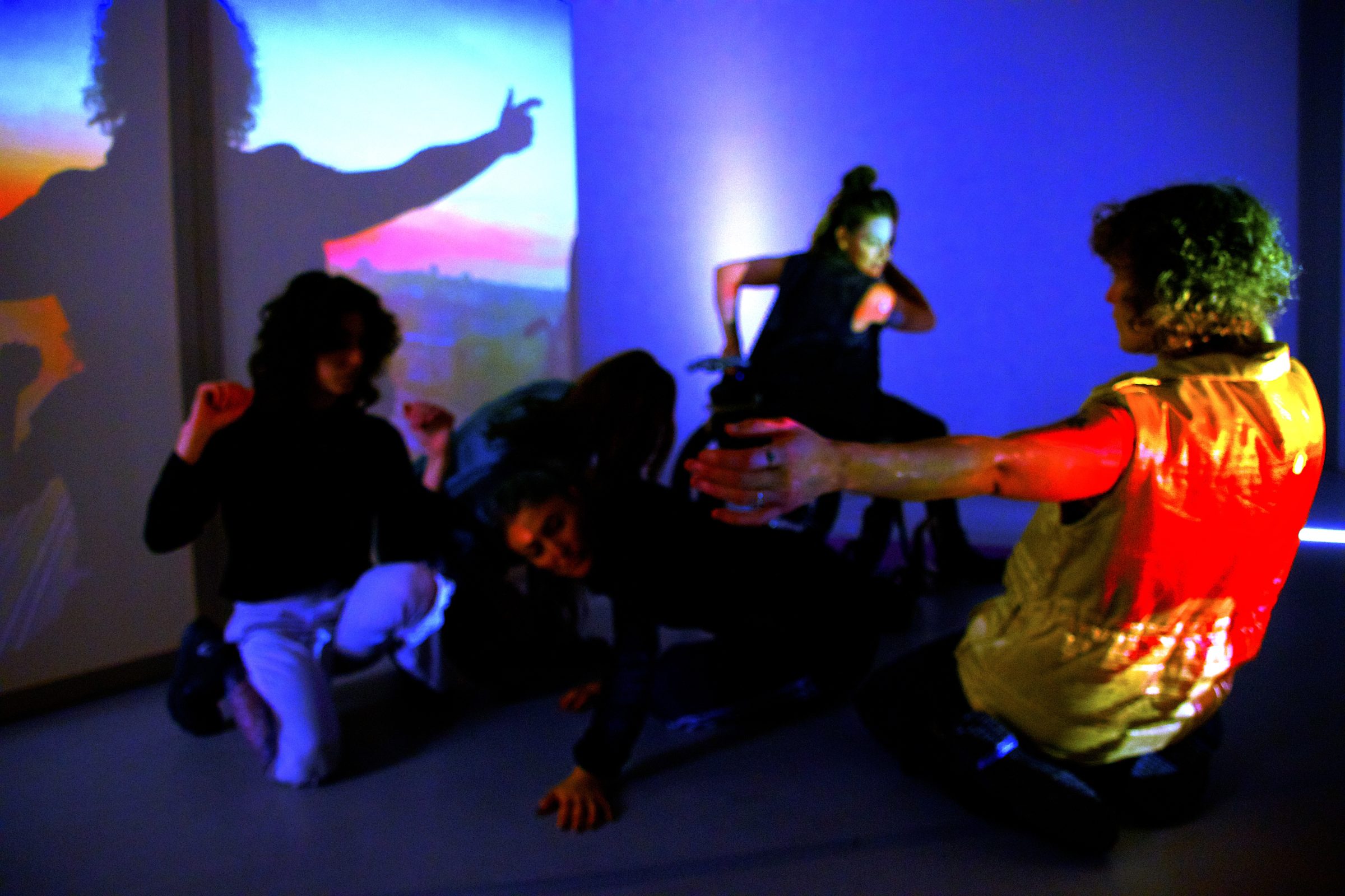 In einem zeitlosen, blau beleuchteten Raum bewegt sich eine Gruppe von fünf weißen Körpern vor einem projizierten Sonnenuntergang. Das Licht spiegelt sich auf ihrer Haut und wirft leuchtend rote, blaue und orangefarbene Streifen. In der Mitte schmelzen zwei Körper in Richtung Boden, verlagern ihr Gewicht und geben sich der Schwerkraft hin. Unten trifft eine weiche, offene Handfläche auf den Boden, trotzt dem Zusammenbruch, der Kopf ist leicht geneigt und ruht auf einem Luftkissen. Zwei umkreisende Körper beziehen ihre Energie aus dem Kern, ihre Präsenz ist aufgeladen. Die Anziehungskraft bestimmt ihre Bewegungen: ein nach oben gebeugter Arm, eine Hand, die sich am Rollstuhl festhält und freudig eine Drehung erwartet; ein weiteres Paar Arme, die sich erhoben haben, weiche, geschlossene Fäuste, die entschlossen in der Luft schweben. Mit dem Gesicht zur Leinwand umarmt eine Person mit lockigem Haar den Raum um die Gruppe herum. Mit ausgestreckten Armen kniet sie zurück, stark und fürsorglich, und dehnt dessen Grenzen aus. Ihr Rücken wird nun zu einer großen roten Sonne, die auf eine goldene, ärmellose Weste aus den 80er Jahren scheint. Ein Schatten fällt auf die gesättigte Projektion und verdoppelt die Sonnenfigur über der Landschaft.