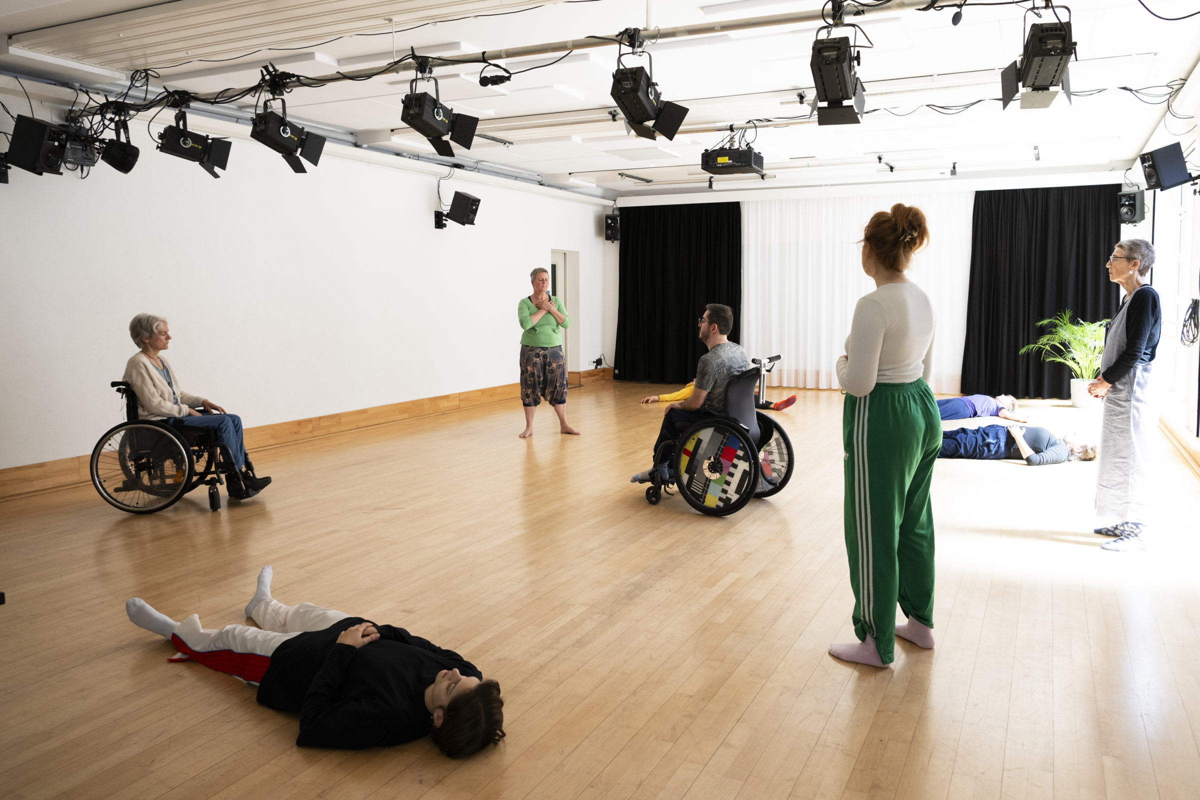 Menschen in Sportkleidung befinden sich verteilt in einem hellen Raum mit Holzboden. Einige Personen liegen auf dem Boden, andere sitzen entspannt in ihren Rollstühlen oder stehen im Raum.