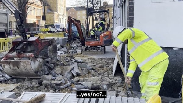 Auf einer Baustelle ist der Gehweg großflächig aufgebrochen. Zwischen Schutthaufen arbeiten mehrere Personen in Warnkleidung. Links bewegt ein großer Baggerarm schwere Betonstücke, weiter hinten bedient jemand einen kleinen Minibagger mit Presslufthammer. Rechts steht eine Person nahe einer Hauswand und stützt sich auf eine Schaufel. Fahrzeuge und Absperrgitter rahmen die Szene in einer belebten Straßenumgebung. Am unteren Bildrand steht eine Bildunterschrift mit der Aufschrift „…ja/nein?…“ in weißer Schrift auf schwarzem Hintergrund.