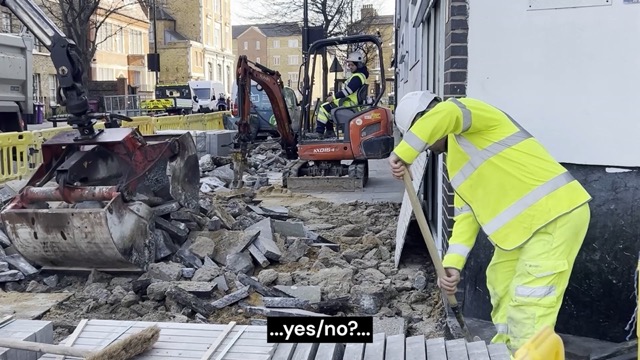 Auf einer Baustelle ist der Gehweg großflächig aufgebrochen. Zwischen Schutthaufen arbeiten mehrere Personen in Warnkleidung. Links bewegt ein großer Baggerarm schwere Betonstücke, weiter hinten bedient jemand einen kleinen Minibagger mit Presslufthammer. Rechts steht eine Person nahe einer Hauswand und stützt sich auf eine Schaufel. Fahrzeuge und Absperrgitter rahmen die Szene in einer belebten Straßenumgebung. Am unteren Bildrand steht eine Bildunterschrift mit der Aufschrift „…ja/nein?…“ in weißer Schrift auf schwarzem Hintergrund.