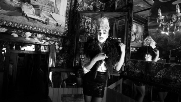 Schwarz-weißes Reenactment einer Karl Lagerfeld-Serie aus den 1980er-Jahren im Berliner Club Kumpelnest 3000. Rafael-Evitan Grombelka übernimmt die Rolle von Gunter Trube und reinszeniert Look und Pose direkt im Originalraum. Mit hochgestecktem Haar, Make-Up, kurzem Rock, Bluse und federartiger Stola nimmt er die Bildsprache der Serie auf. Spiegel, gemusterte Wände und ein Kronleuchter gliedern den Raum, im Hintergrund ist ein Wandbild zu sehen, das ein Kind zeigt, das an einen Löwen angelehnt ist.