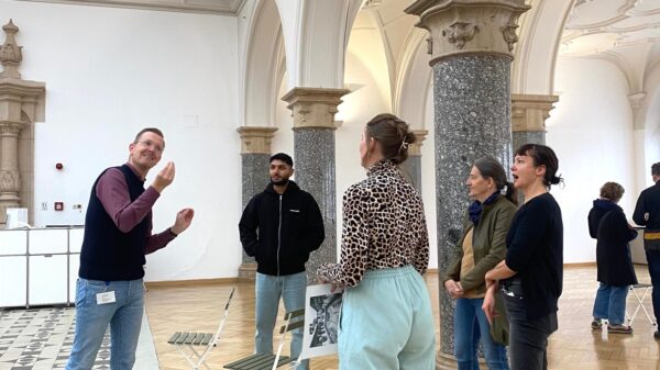 Foto eines Kunstgesprächs in der Rathausgalerie: Der Museums Signer beschreibt in DGS - Deutscher Gebärdensprache die Ausstellung für vier Besucher*innen, die im Halbkreis um in herum stehen.