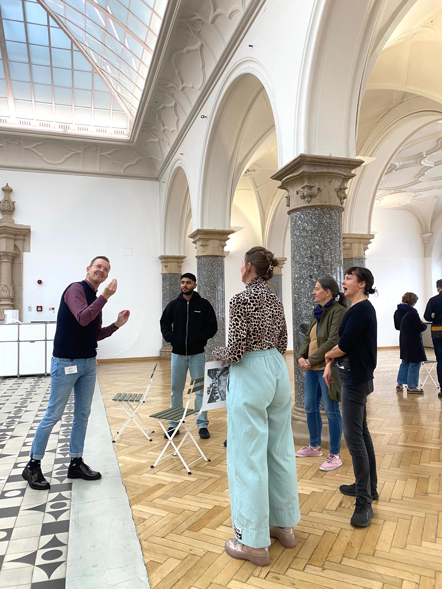 Foto eines Kunstgesprächs in der Rathausgalerie: Der Museums Signer beschreibt in DGS - Deutscher Gebärdensprache die Ausstellung für vier Besucher*innen, die im Halbkreis um in herum stehen.