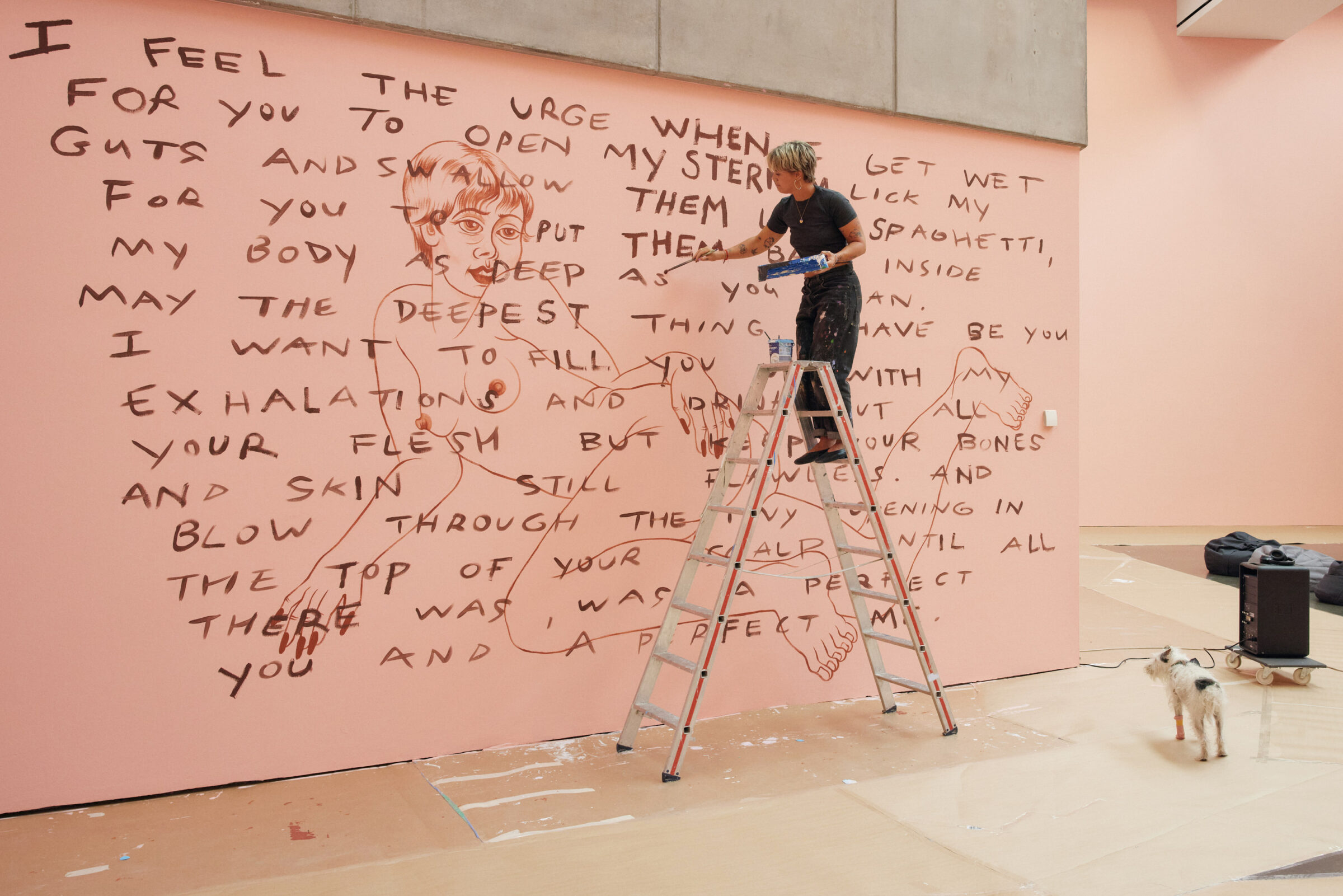 Eine junge, blonde Frau steht auf einer Leiter und beschreibt eine rosa-gestrichene Wand mit einem Text.