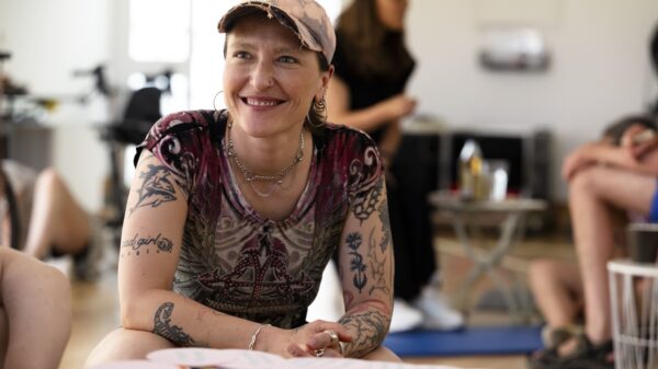Referent*in Agnieszka Habraschka kniet im Un-Label Studio auf dem Boden. Agnieszka ist weiß, ihre Arme und Hals sind tätowiert und they trägt eine ausgefranste Cap. Agnieszka lächelt jemanden an. Im Hitergrund sind workshopteilnehmer*innen zu erkennen.