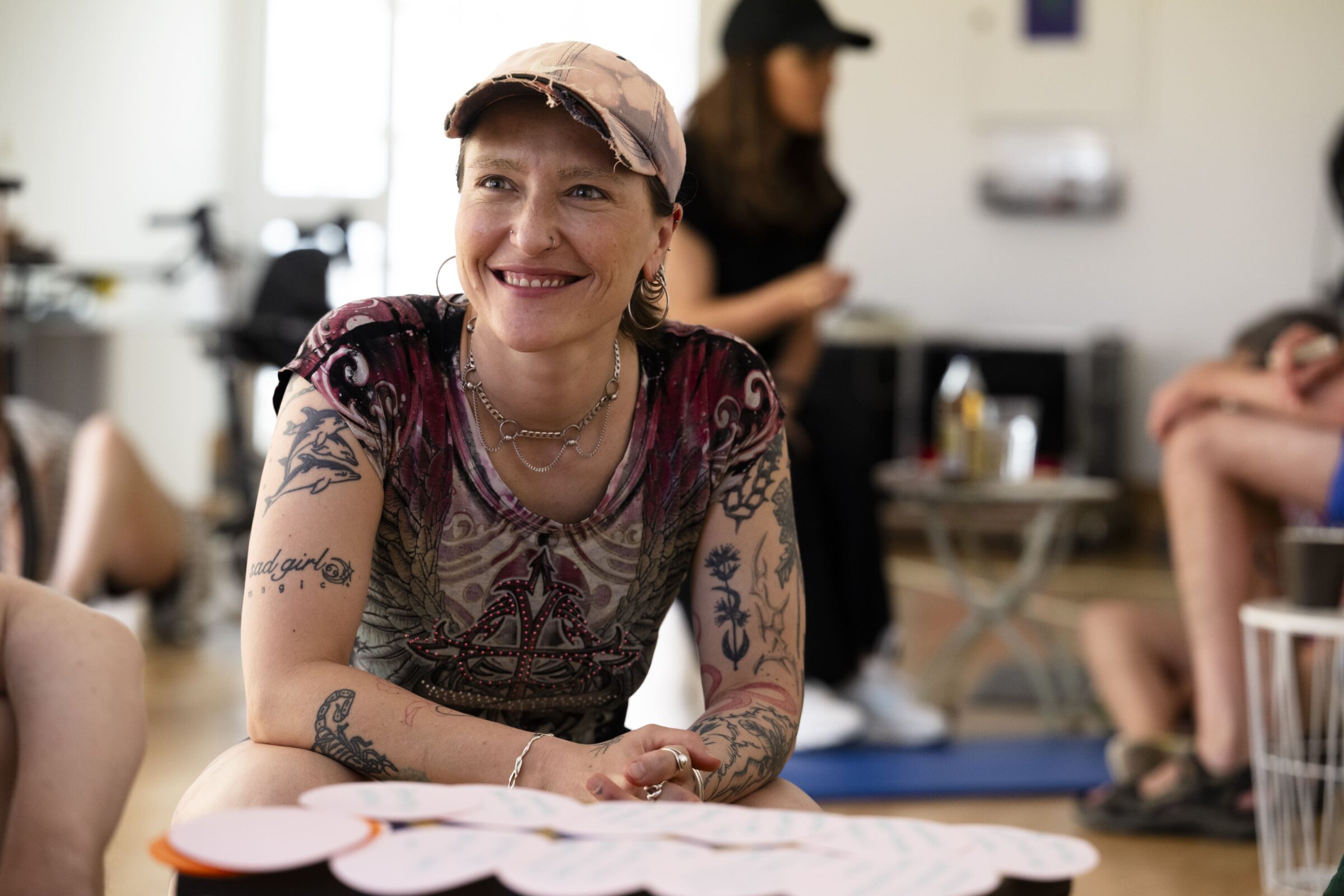 Referent*in Agnieszka Habraschka kniet im Un-Label Studio auf dem Boden. Agnieszka ist weiß, ihre Arme und Hals sind tätowiert und they trägt eine ausgefranste Cap. Agnieszka lächelt jemanden an. Im Hitergrund sind workshopteilnehmer*innen zu erkennen.