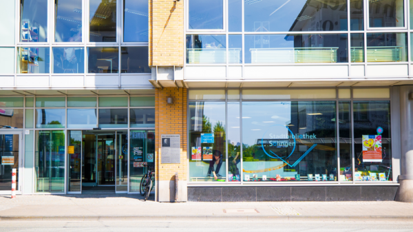 Außenansicht eines Gebäudes mit großen Glasfenstern und Eingangstüren. In der rechten Fensterfront steht: „Stadtbibliothek Solingen“. In der Mitte an der Wand hängt eine Tafel mit der Überschrift „EMIL KRONENBERG-HAUS“. Im Schaufenster liegen Bücher aus. Ein Fahrrad steht vor dem Eingang.